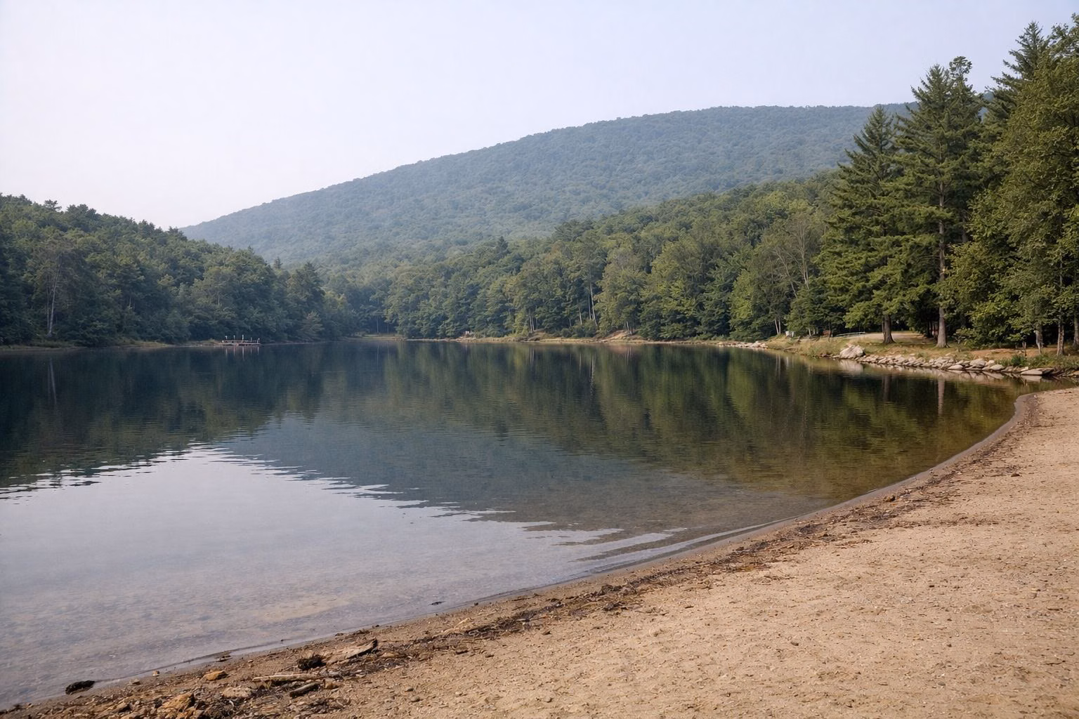 A calm small mountain lake with a narrow sand beach in the foreground and forested ridges behind, no people